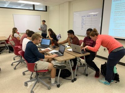 The image shows several students at one table working on their laptop computers with Mary Lofton assisting one student.